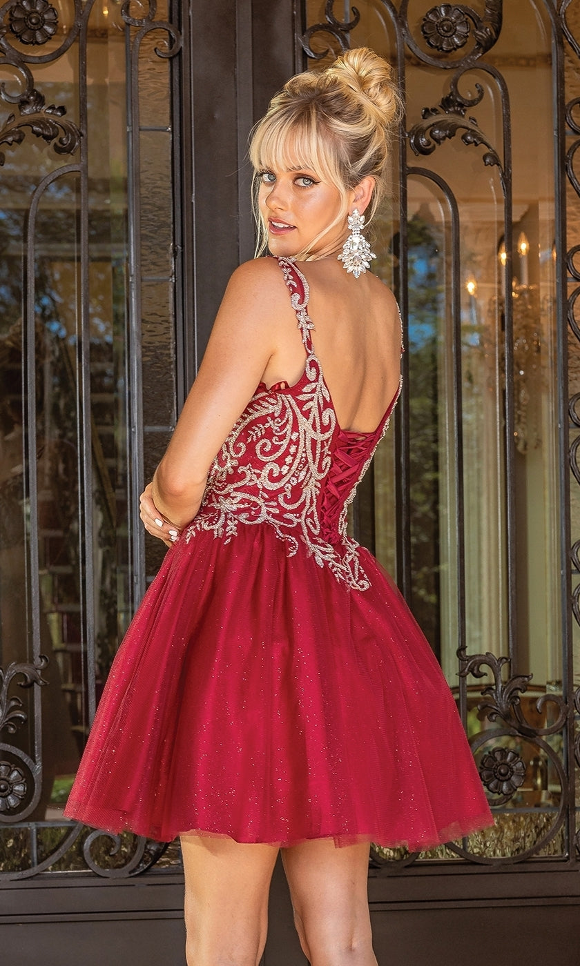 A woman in the Embroidered-Bodice Short Glitter Hoco Dress 3295 with silver details and statement earrings stands before ornate black doors, glancing back over her shoulder.